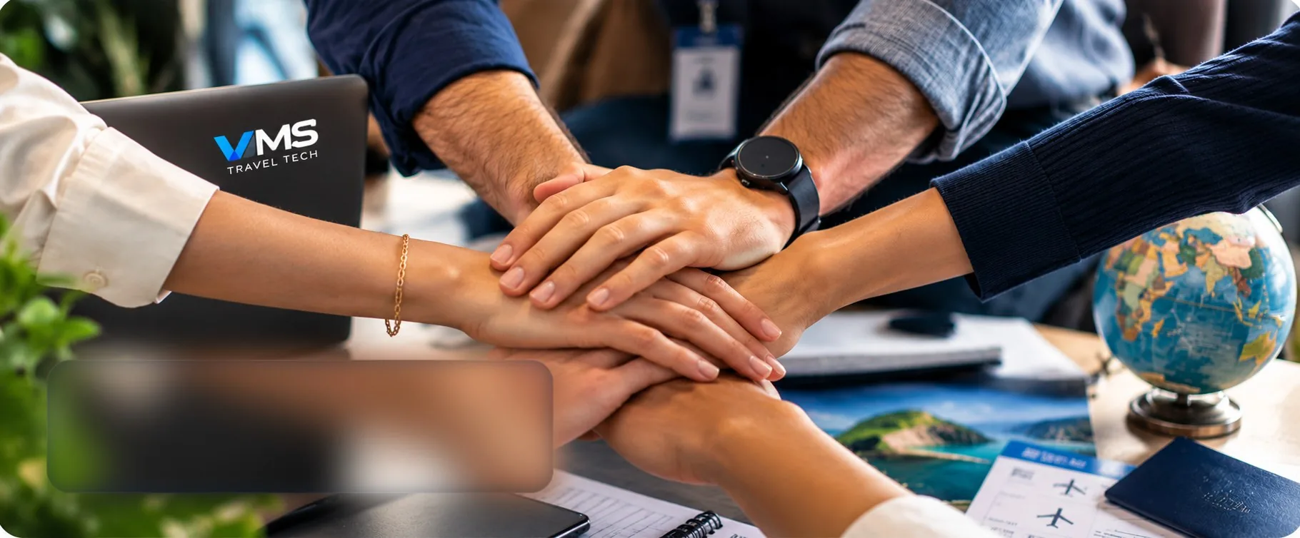 VMS colleagues joining hands over a desk with a globe, tickets and a VMS laptop
